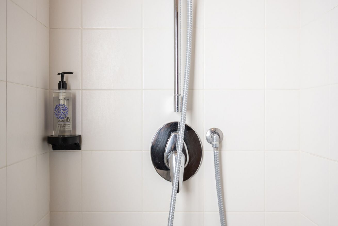 Ducha con cabezal de ducha de mano y grifo empotrado en la pared, dispensador de jabón en un estante y paredes de azulejos blancos.