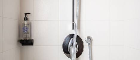 Ducha con cabezal de ducha de mano y grifo empotrado en la pared, dispensador de jabón en un estante y paredes de azulejos blancos.
