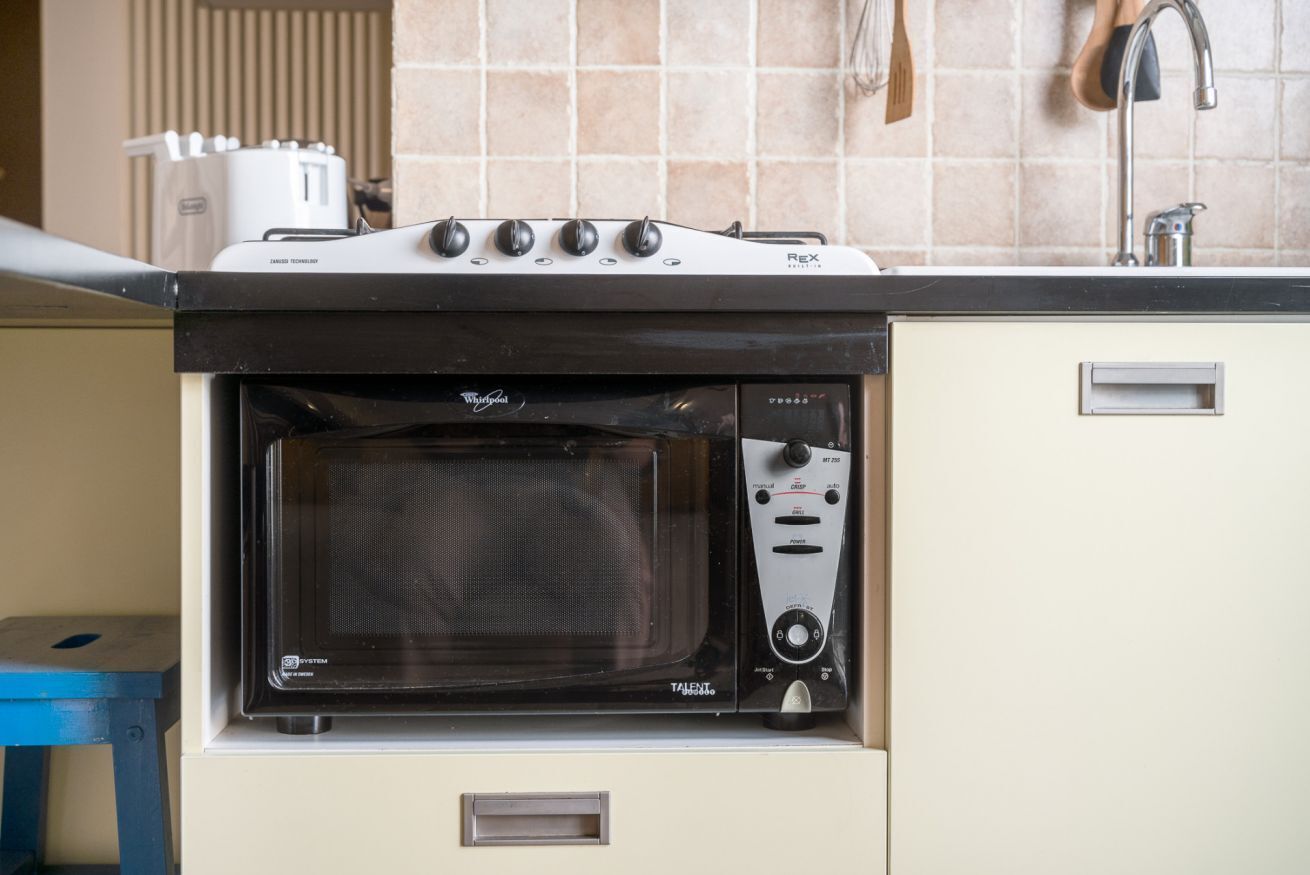 Uma cozinha compacta com micro-ondas e forno embutidos, uma pia e um banquinho azul.