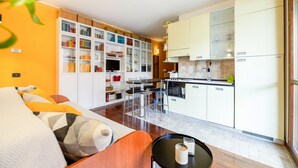 Una sala de estar moderna con un sofá, una mesa de comedor y una zona de cocina con armarios blancos y un horno empotrado.