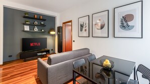 Un salon moderne avec un canapé gris, une table à manger en verre et un téléviseur affichant le logo « NETFLIX ». Des œuvres d’art encadrées ornent le mur, et une étagère présente divers objets décoratifs.