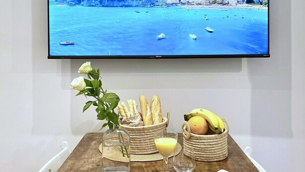 Ein Esstisch mit Croissants, Brot, Bananen, einem Glas Orangensaft und einer Weinflasche, mit einem Fernseher, der eine Küstenlandschaft zeigt.