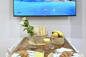Une table à manger avec des croissants, du pain, des bananes, un verre de jus d’orange et une bouteille de vin, devant une télévision qui diffuse une scène de paysage côtier.