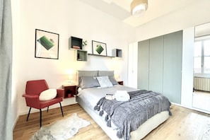 Um quarto com cama, uma cadeira vermelha, mesas de cabeceira, guarda-roupa e quadros nas paredes.