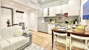 Eine moderne Küche mit weißen Schränken, einem Esstisch mit Stühlen und einem Wohnbereich mit Sofa und Pflanze.