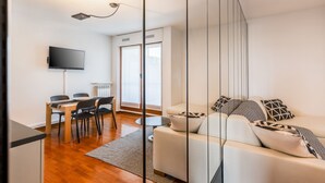Un salon moderne comprenant un canapé, un téléviseur à écran plat, un coin repas avec des chaises et une table, ainsi qu’une fenêtre avec des stores.