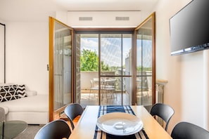 Un salon moderne comprenant un canapé, une table à manger avec des chaises, un téléviseur fixé au mur et une porte-fenêtre coulissante donnant sur une terrasse extérieure.