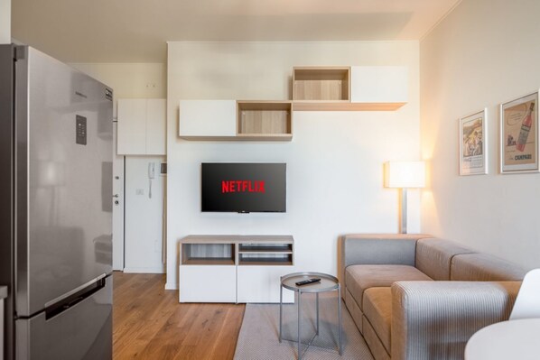Ein modernes Wohnzimmer mit Flachbildfernseher, einer Couch, einem Couchtisch und einer Küchenzeile mit Kühlschrank.