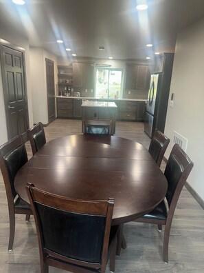 A dining area with a round table, six chairs, and a kitchen in the background.