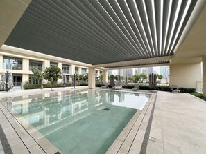 Un espace piscine intérieur moderne, doté d’une structure en verre, de chaises longues et d’un coin salon couvert avec des parasols.