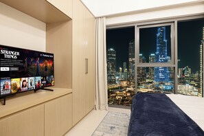 Une chambre d’hôtel avec une grande télévision diffusant un film, une vue sur la ville par la fenêtre et un lit recouvert d’une couverture bleue.