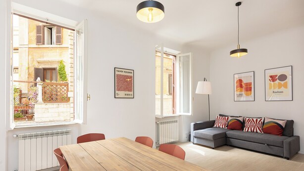 A modern living room with a wooden dining table, a grey sofa, and a view of a building through an open window. - Rental in Rome Mastai Trastevere Home (Rome)