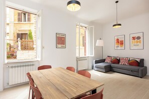 A modern living room with a wooden dining table, a grey sofa, and a view of a building through an open window. - Rental in Rome Mastai Trastevere Home (Rome)
