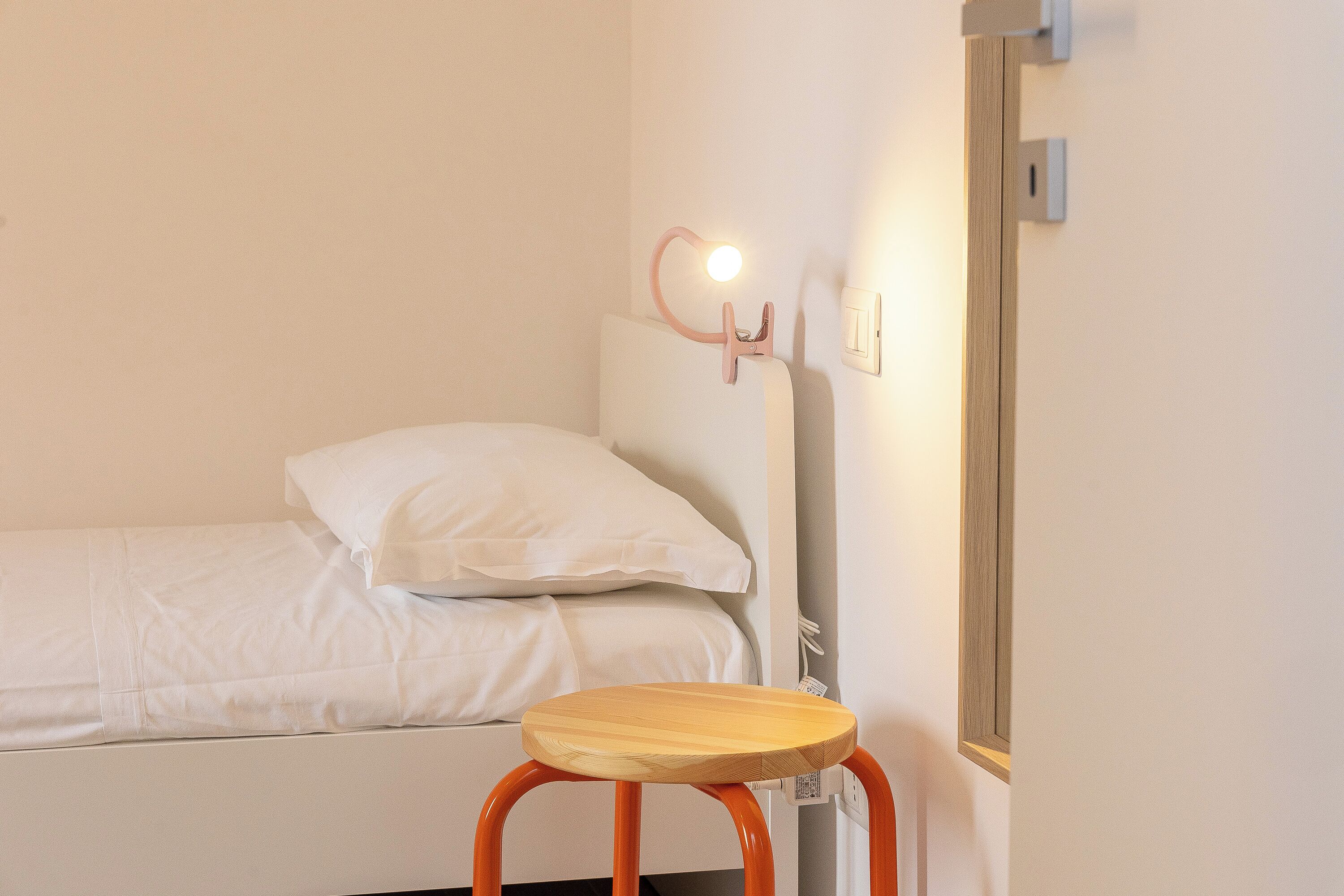 Una cama individual con ropa blanca, un taburete de madera y una lámpara fijada a la pared.