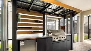 An outdoor kitchen with a built-in grill, a stainless steel refrigerator, and a window overlooking a garden. - 7 Bd / 6.5 Ba / Sleeps 20 / Solara Resort (Kissimmee)