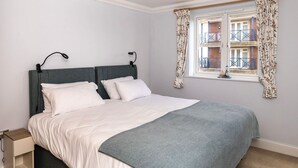A neatly made bed with a grey headboard, white bedding, and a grey throw blanket. There is a window with floral curtains and a view of a balcony. - Harbour Hideaway - great views & flexible beds (Eastbourne)