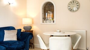A living room with a blue sofa, a round table with two chairs, a lamp, a clock, and a window with a view of the beach. - Harbour Hideaway - great views & flexible beds (Eastbourne)
