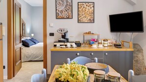 Une chambre moderne comprenant un lit, un coin repas avec une table et des chaises, un comptoir de cuisine avec divers ustensiles, et un téléviseur fixé au mur.