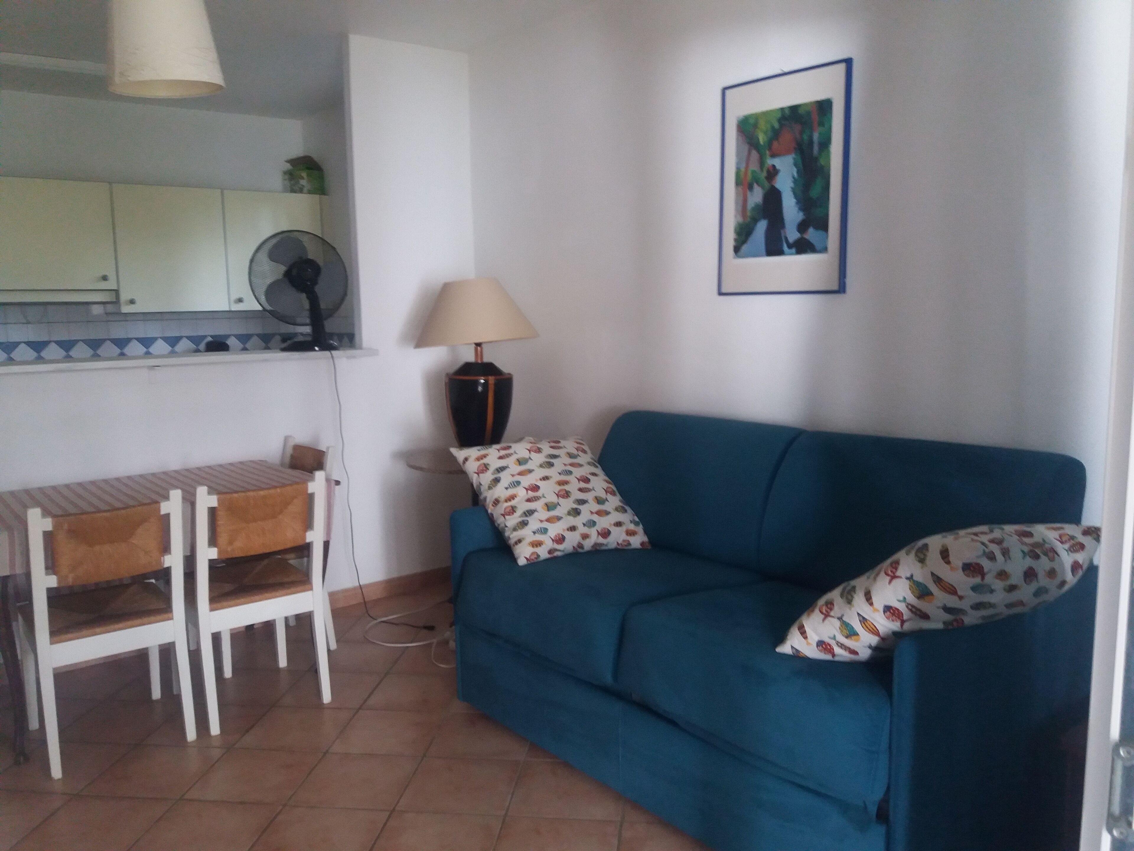 Ein Wohnzimmer mit blauem Sofa, einem kleinen Esstisch mit Stühlen, einer Lampe und einem Bild an der Wand.