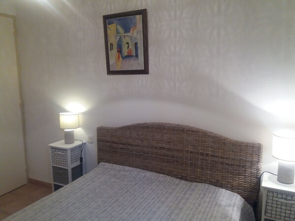 A bedroom with a bed, two bedside tables with lamps, a wicker headboard, and a painting on the wall.