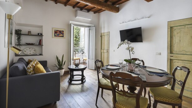 Een gezellige woonkamer met een bank, een eettafel gedekt voor twee personen en een aan de muur bevestigde televisie.