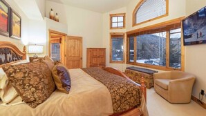 Een slaapkamer met een groot bed, een houten bankje, een stoel en een raam met uitzicht op bomen en sneeuw.