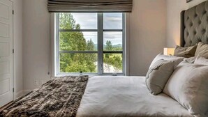 Un dormitorio con un ventanal grande, una cama con ropa de cama blanca y un cabecero gris.