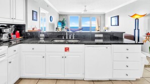 Une cuisine moderne avec des armoires blanches, un comptoir noir et un grand îlot central. On peut admirer l’océan à travers les portes-fenêtres coulissantes.