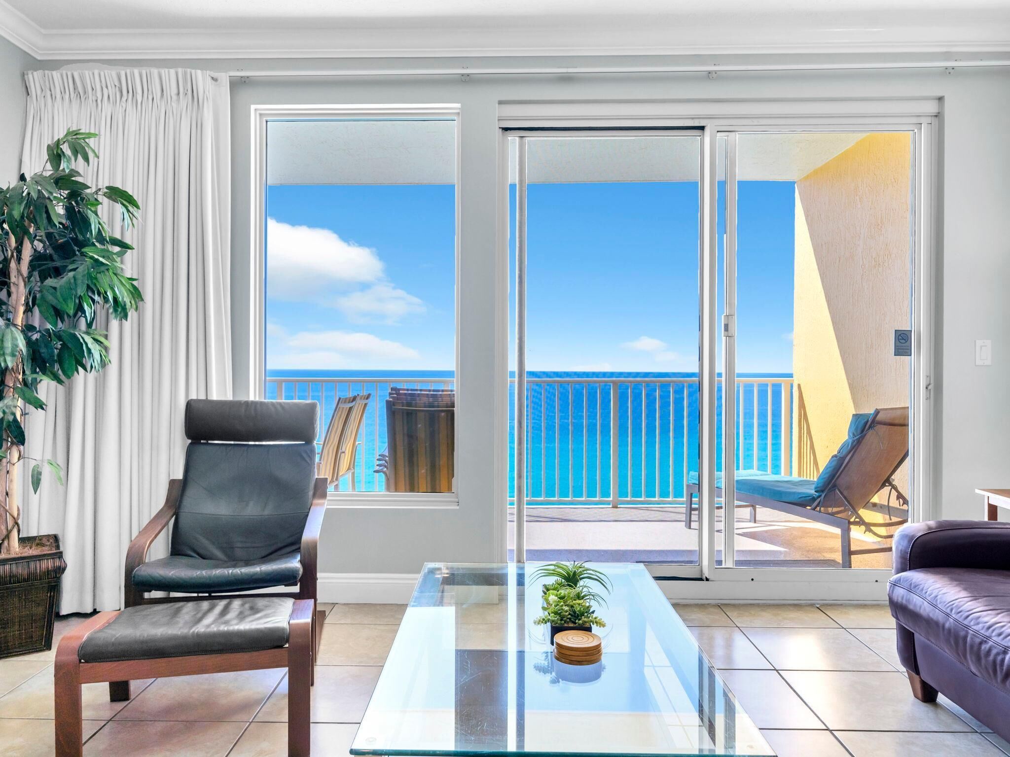 A balcony with a clear view of the ocean, a glass table with a plant, a leather chair, and a sofa.