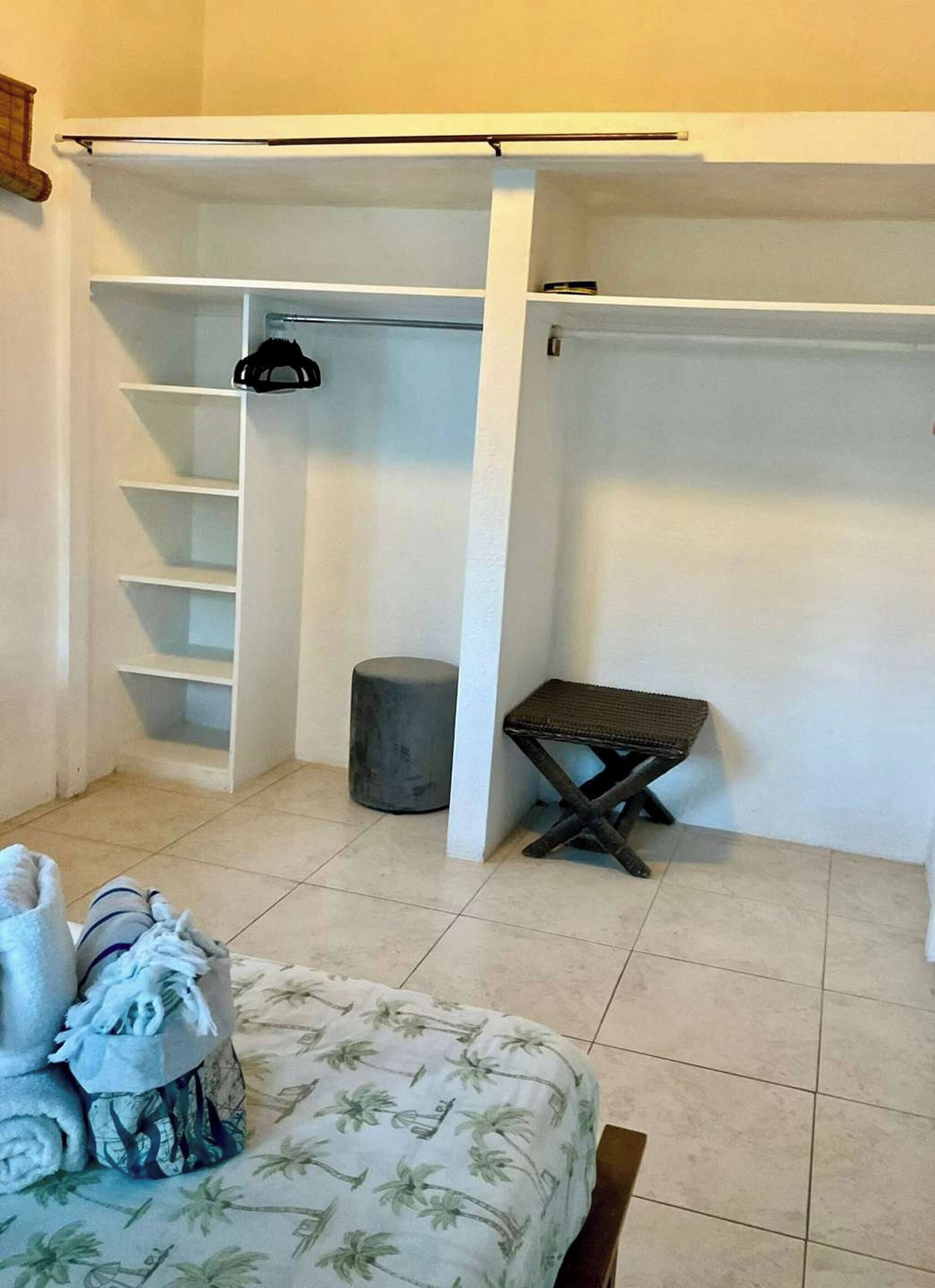A bedroom with a walk-in closet, a grey ottoman, a small bench, and a bed with a floral bedspread.