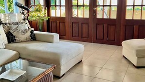 A spacious living room with a large sofa, a glass coffee table, and a wicker side table. There are two large wooden doors leading to an outdoor area. - Harbour View - by Zenbreak (Lodge Hill)