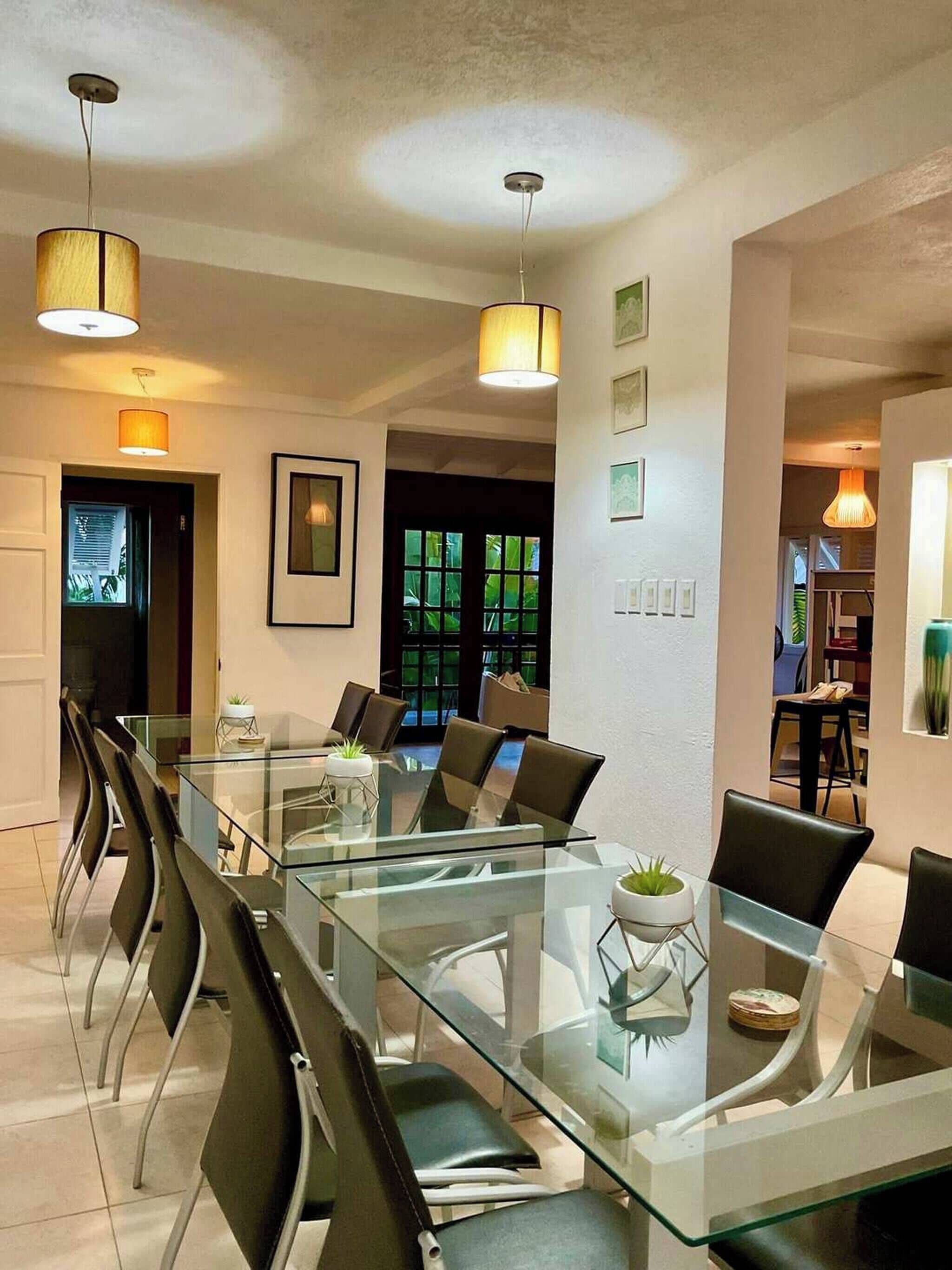 A modern dining area with a glass table and chairs, pendant lighting, and a view of the outdoors.