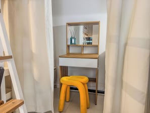 Une pièce avec un bureau en bois, un tabouret jaune, un miroir et un rideau blanc.