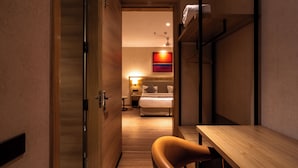 A hotel room with a wooden floor, a bed with white bedding, a desk with a chair, and a shelf with a towel. - Hotel Bluestone Prime Nehru Place by The Cosy hotels (New Delhi)