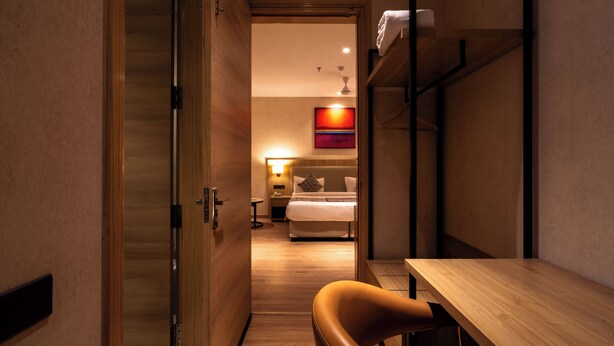 Ein Hotelzimmer mit Holzboden, einem Bett mit weißer Bettwäsche, einem Schreibtisch mit Stuhl und einem Regal mit Handtuch.