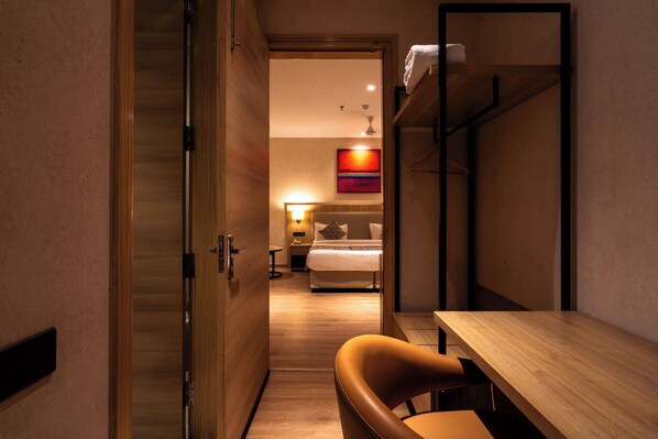 A hotel room with a wooden floor, a bed with white bedding, a desk with a chair, and a shelf with a towel.