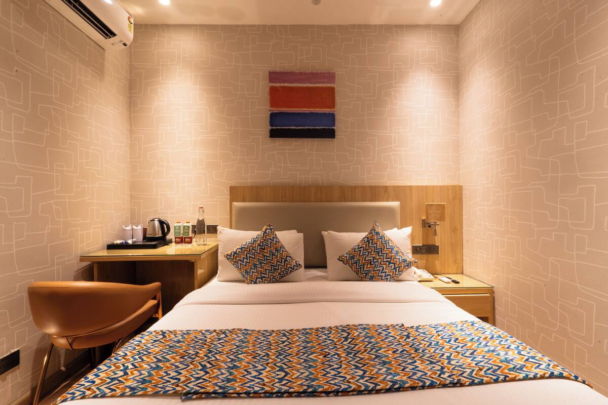 A hotel room with a bed, a desk with a coffee maker, a chair, and a wall-mounted air conditioner.