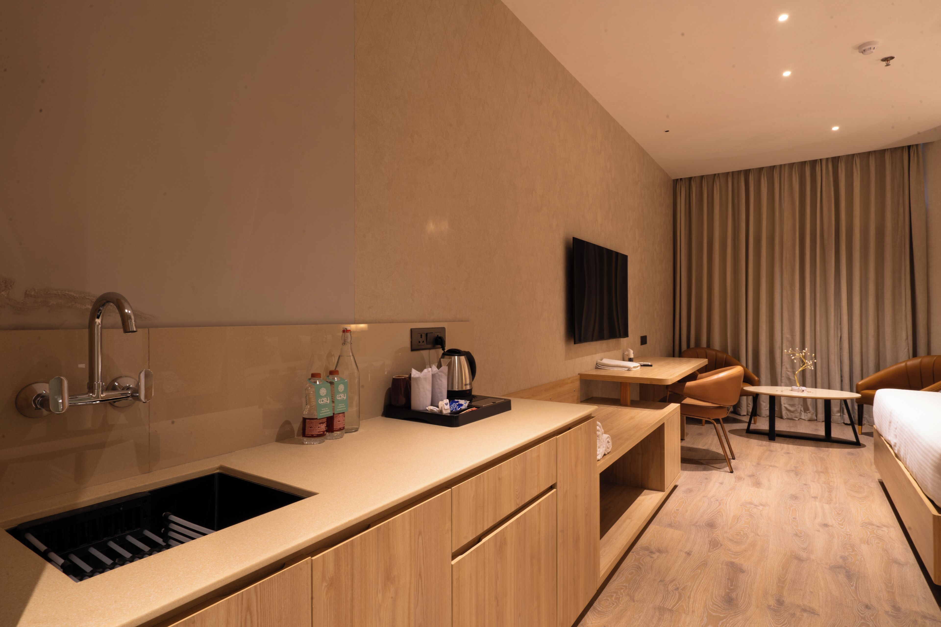 A modern hotel room with a built-in kitchen, a flat-screen TV, a dining area, and a bed.