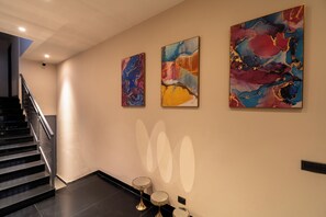 Un couloir avec un escalier, trois tableaux abstraits accrochés au mur et deux petites tables avec des lampes.