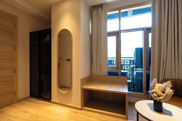 A room with a wooden floor, a bench, a mirror, a window with curtains, and a view of the outside. - Hotel Bluestone Prime Nehru Place by The Cosy hotels (New Delhi)