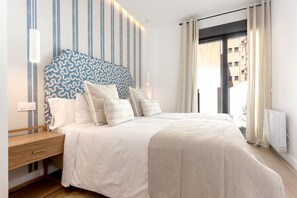 Une chambre à coucher avec un lit, des tables de chevet, une tête de lit décorée et une vue sur des immeubles par la fenêtre.