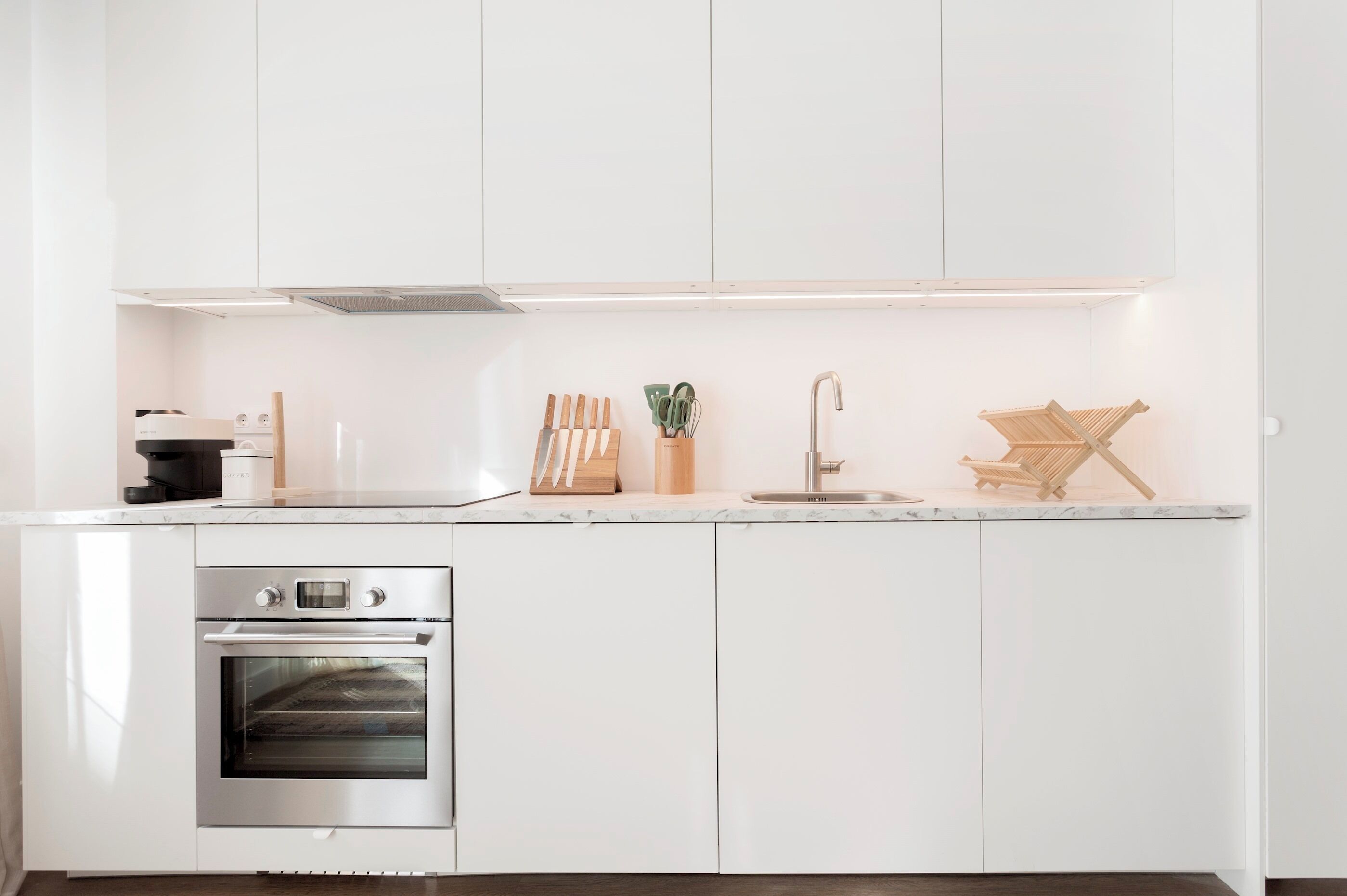 Una cocina moderna con armarios blancos, un horno de acero inoxidable, un lavamanos y utensilios de madera sobre la encimera.