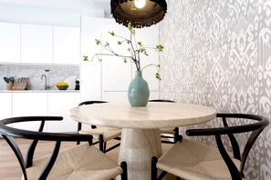 Una mesa redonda con un jarrón de plantas verdes, rodeada de sillas tejidas, en una cocina moderna con armarios blancos y un papel tapiz estampado.