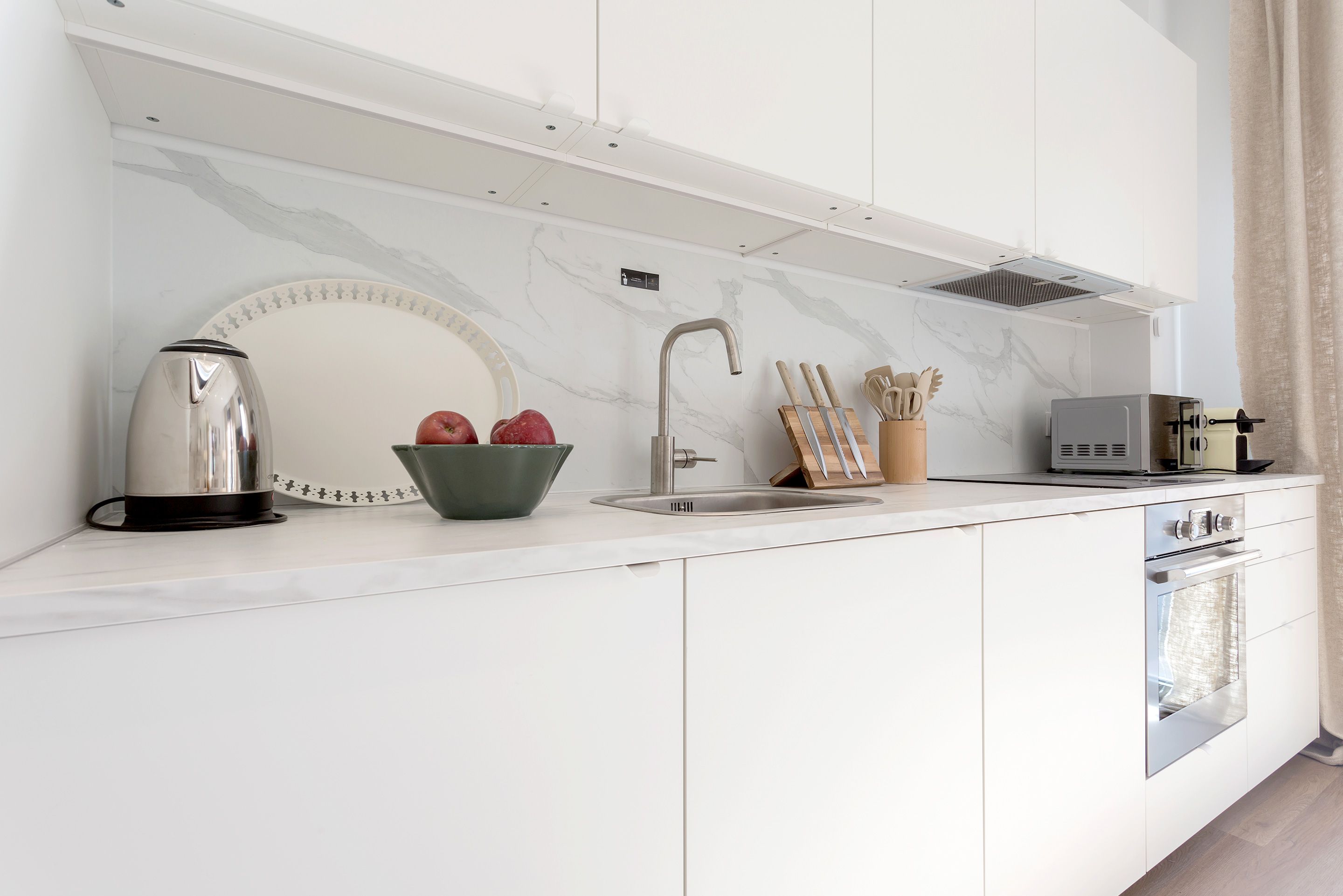 A modern kitchen with white cabinetry, a stainless steel kettle, a bowl of apples, a knife block, and a toaster.