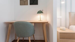 A room with a wooden desk, a green chair, a green desk lamp, and a bedside table with a white container.