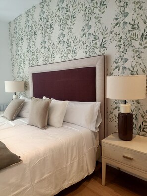 Un dormitorio con cama, almohadas, una mesita de noche, una lámpara y un papel tapiz con motivos florales.