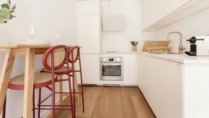 Een kleine keuken met witte kasten, een inbouwoven, een spoelbak en een eethoek met een houten tafel en rode stoelen.