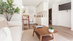 Een moderne woonkamer met een houten salontafel, een bank en een grote plant. In de achtergrond bevindt zich een keuken met witte kasten en een eettafel.