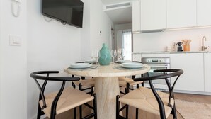 Et spiseområde med et rundt bord, fire stole og en teal vase. Et køkken er synligt i baggrunden med hvide skabe og rustfrie stålgadgetter.