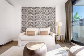 Ein modernes Wohnzimmer mit strukturiertem Wandpapier, einer Couch mit Kissen, einem Beistelltisch mit einer Lampe und Blick ins Freie durch ein Fenster.